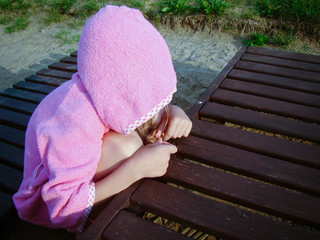 Child is squatting with a hood. Pink clothes. Empty beach chairs, sand. Concept of loneliness, emotional experiences.