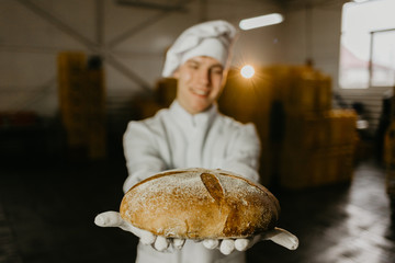 Young happy female worker in sterile cloths holding freshly baked cookies on tinplate inside food...