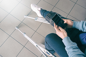 Woman on crutches, leg is splinted, siting in waiting chair in hospital and using smartphone. Knee brace support, bandaged leg cast after injury accident. Rehabilitation, travel insurance concept.