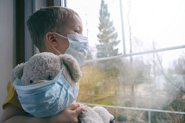 Sad illness child on home quarantine. Boy and his teddy bear both in protective medical masks sits...