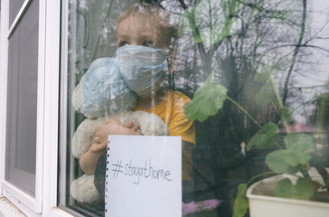 Stay at home quarantine for coronavirus pandemic prevention. Child and his teddy bear both in protective medical masks sits on windowsill and looks out window. View from street. Prevention epidemic.