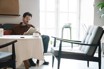 Image of handsome unshaven young man in eyeglasses reading book in cafe