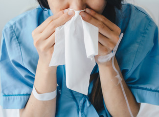 Fototapeta premium Cropped shot view of young woman wearing patient cloths, sneezing in tissue at hospital. Conceptual of sickness woman feeling when admitted in the hospital.