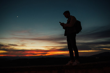 Silhouette of young man with mobile phone in beautiful sunset. Young brave man standing on the edge...