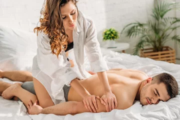 Sinnliches Mädchen im weißen Hemd, das dem sexy Freund, der mit geschlossenen Augen auf dem Bett liegt, eine erotische Armmassage macht © LIGHTFIELD STUDIOS
