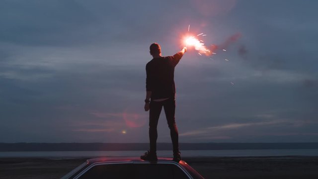 Handheld cinematic dark shot of young man standing on the car with red signal burning flare on the beach near the water