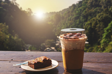 Iced latte with milk foam on top, and chocolate brownie in a white dish on a wooden table. Natural hill is a background. Nice holiday. Another drink menu in summer. Close-up photo.