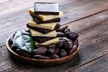 Stack of dark bitter chocolate and cocoa butter with whole cacao beans in wooden bowl original...
