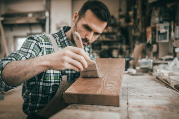 Craftsman applies varnish on wooden board by paintbrush in his carpentry workshop