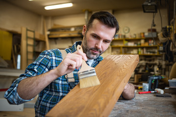 Craftsman applies varnish on wooden board by paintbrush in his carpentry workshop