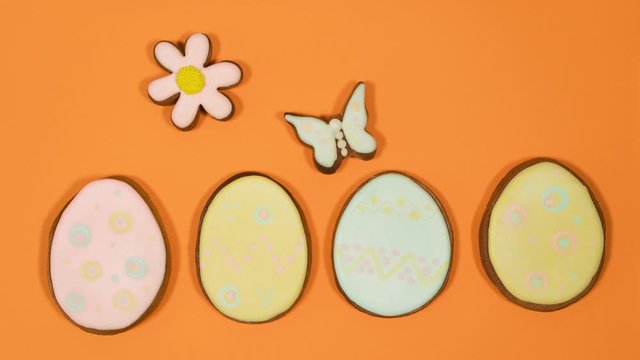 Closeup view of sweet colorful ginger cookies in shapes of Easter eggs, butterfly, bird and flower moving cheerfully on bright orange background. Animated cute holiday video background in 4k.