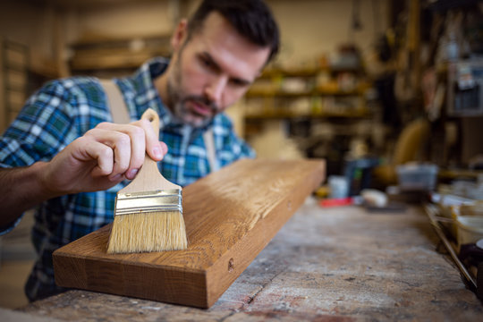 Craftsman applies varnish on wooden board by paintbrush in his carpentry workshop