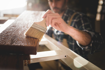 Close up of paintbrush applies paint or varnish on wooden board in carpentry workshop