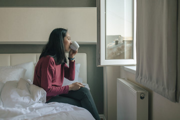 Woman is drinking hot coffee and looking out to the window in her apartment.