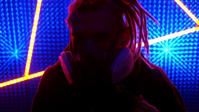 Young guy model with dreadlocks in neon lights. Neon triangle in a club studio. Concept of dubstep or punk style. Wearing a gas mask from dirty air. Protected in a public place from the coronavirus