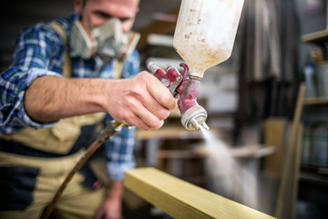 Carpenter with paint spray gun painting wooden plank in carpentry workshop