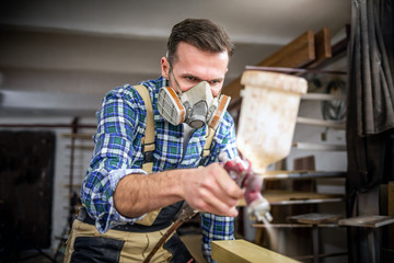 Carpenter with mask using paint spray gun to painting wooden plank in carpentry workshop