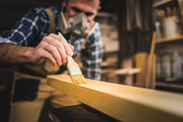 Carpenter with mask applies paint using paintbrush in carpentry workshop