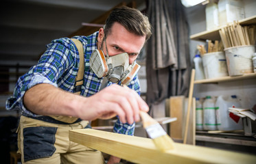 Fototapeta na wymiar Carpenter with mask applies paint using paintbrush in carpentry workshop