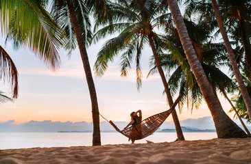 Young woman sitting in hammock swinging on the exotic island sand beach at sunrise time