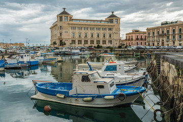 Fishing and sailing boats in the marina of Ortygia island in Sicily at sunrise with cloudy sky 