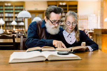 Handsome man grandfather reading a book for his cute granddaughter, hugging her while sitting together at the table in vintage old library.