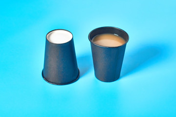 One full and inverted black cardboard glass for hot beverages on blue background. Concept of environmental protection, coffee break, ecology, garbage recycle