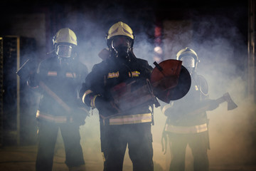 Fototapeta premium Group of professional firefighters wearing full equipment, oxygen masks, and emergency rescue tools, circular hydraulic and gas saw, axe, and sledge hammer. smoke and fire trucks in the background.