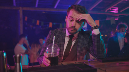 Fototapeta na wymiar Handsome tired businessman sitting at bar counter drinking alcohol thinking over life with upset expression at night youth party in the club.