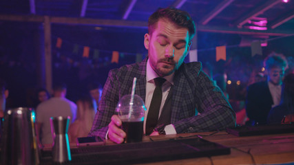 Modern businessman browsing financial news on smartphone drinking at bar counter trying to relax at the party. Night life.