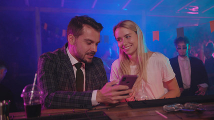 Pretty blondie and her boyfriend businessman laughing of jokes looking on mobile phone leaning on bar counter in modern club at night.