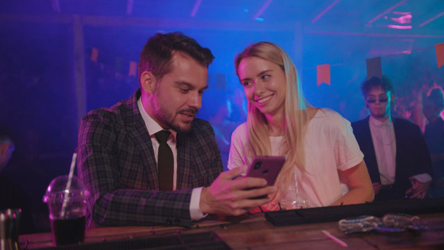 Pretty Blondie And Her Boyfriend Businessman Laughing Of Jokes Looking On Mobile Phone Leaning On Bar Counter In Modern Club At Night.