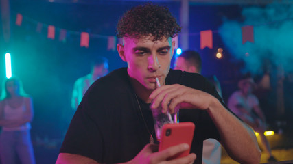 Close-up of handsome caucasian boy with black curly hair drinking cocktail using smartphone social...