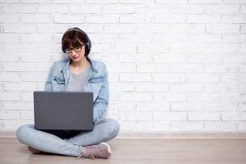 young woman or teenage girl sitting on the floor, using laptop and listening music in headphones over white brick wall background