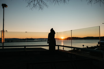 Fototapeta premium Silhouette of young European Muslim woman with hijab holding mobile phone and taking photo of beautiful sunset in Split. 
