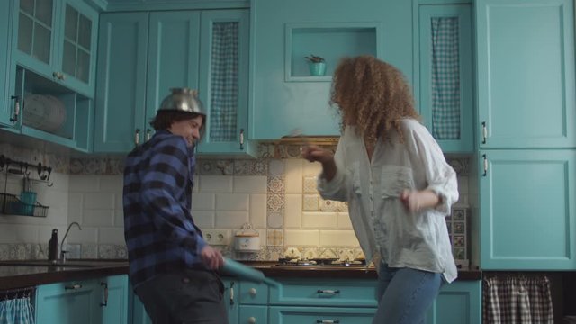 Young happy 20s couple in casual clothing having fun with kitchenware in hands on cozy blue kitchen. Man and curly woman funny fighting with rolling pin, colander and scoop at home kitchen