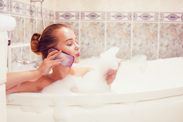 Obraz premium The girl is lying in the whirlpool with foam and talking on a waterproof phone. The girl relaxes and gossips. Poetz girl in the foam. Conversation with a girlfriend.