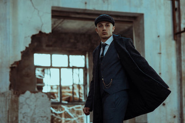 A man posing in the image of an English retro gangster dressed in a coat, suit and flat cap.