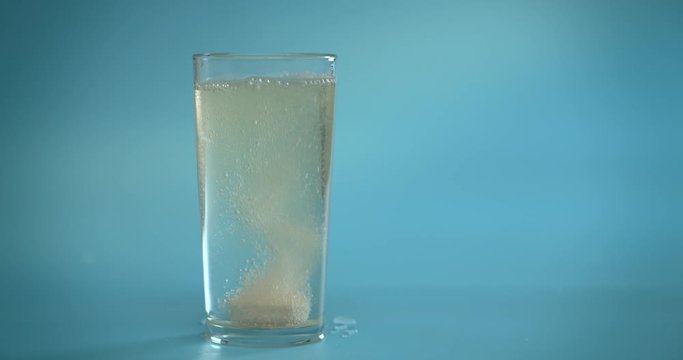A vitamin C tablet is thrown into a transparent glass glass with water. The effervescent pink tablet dissolves. Glass on a blue background. Slow motion