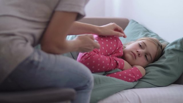 Mother is giving digital thermometer to her cold little daughter, lying on a couch in home