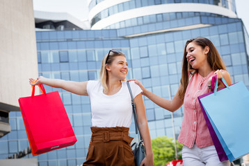 Two smiling young woman walking down the street after sopping, carrying bags in their hands.