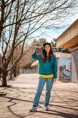 Fototapeta premium A young Caucasian hipster woman poses with a skateboard over her shoulders. In the background, an alley. Vertical. Concept of sports lifestyle and street culture