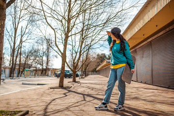 A young Caucasian hipster woman stands with a skateboard in her hand and looks into the distance. In the background, an alley. Concept of sports lifestyle and street culture