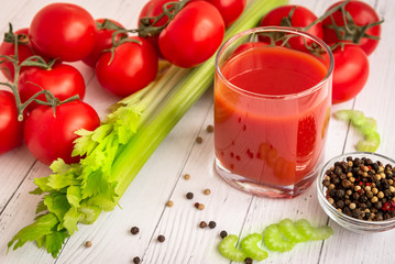 Tomato juice in a glass glass , celery stalks , pepper mix, tomatoes on a branch in the background on a white wooden table.