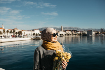 portrait of European Muslim women with hijab sitting on the stone beach with sea and port in the background. She is holding bouquet of yellow flowers.