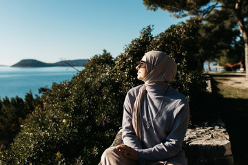 Fototapeta premium portrait of young European Muslim women with hijab sitting on the stone beach with sea and port in the background. She is happy and enjoying sun.