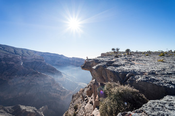 Man peeking over the cliff with the sun in the background at the Grand Canyon of Jebel Shams, Oman - obrazy, fototapety, plakaty