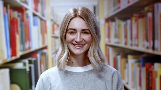 Close-up of amazing girl young student smiling happy at camera staying at library bookshelf background studying. Beautiful female portrait.