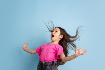 Astonished, flying hair. Caucasian little girl's portrait on blue studio background. Beautiful female model in pink shirt. Concept of human emotions, facial expression, sales, ad, youth, childhood.