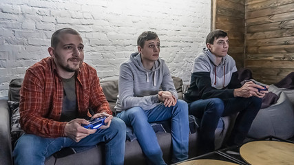 Two guys playing on the console sitting on the couch, one man watching the game. Portrait of three excited handsome men playing video game with console controller in hands